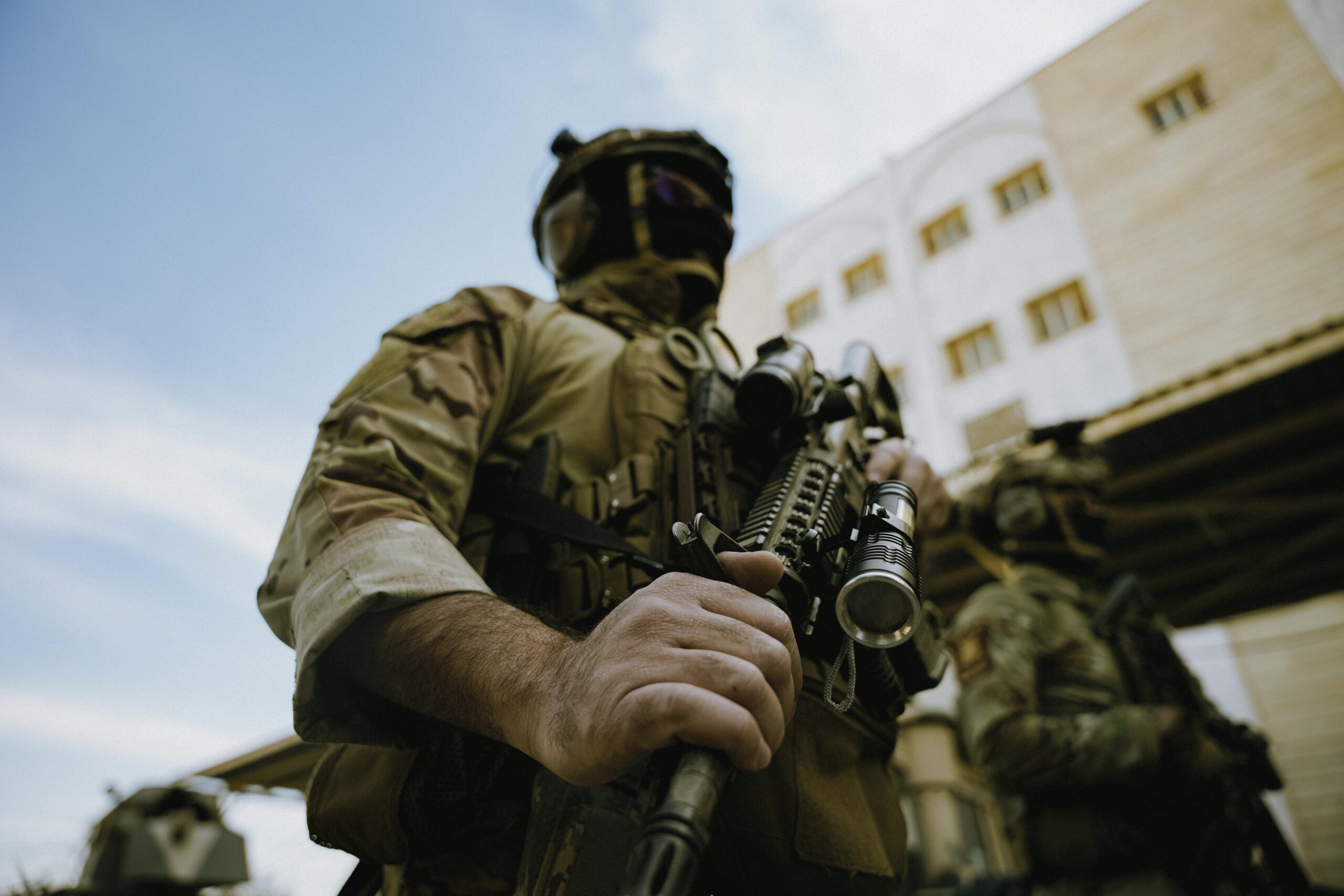 A soldier in tactical gear holding a rifle, outdoors by buildings.